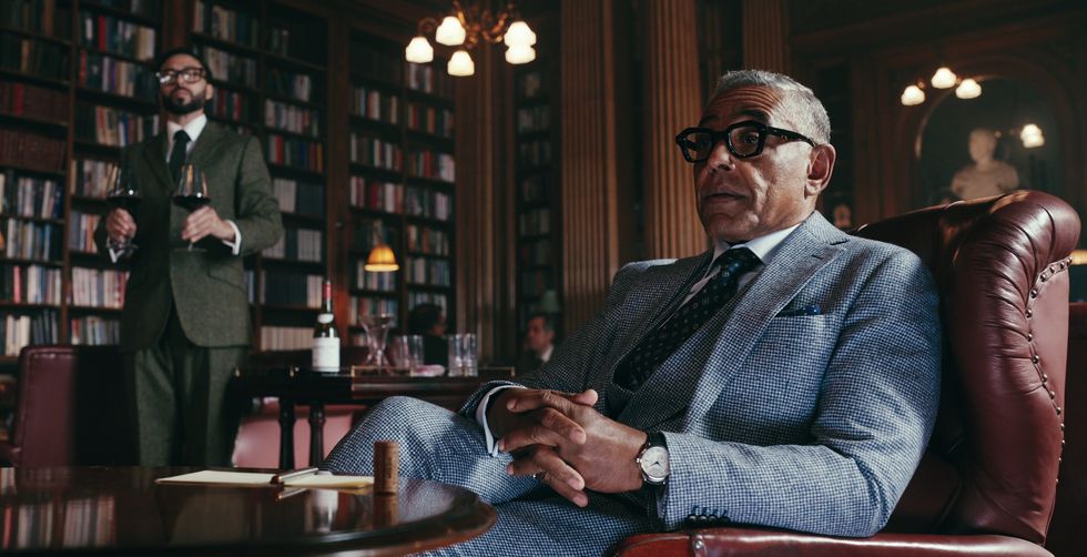 giancarlo esposito dans les messieurs, un homme aux cheveux grisonnants et aux lunettes est assis dans un fauteuil dans une pièce avec des livres bordant le mur, un autre homme s'approche de lui en tenant deux verres de vin