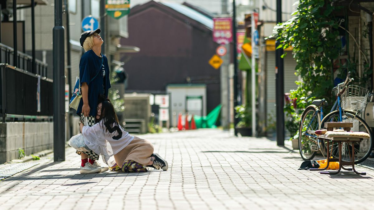 Akari Takaishi serre Saori Izawa agacée autour de ses jambes, alors qu'Izawa se tient au milieu d'un chemin pavé, dans Baby Assassins 2 Babies.