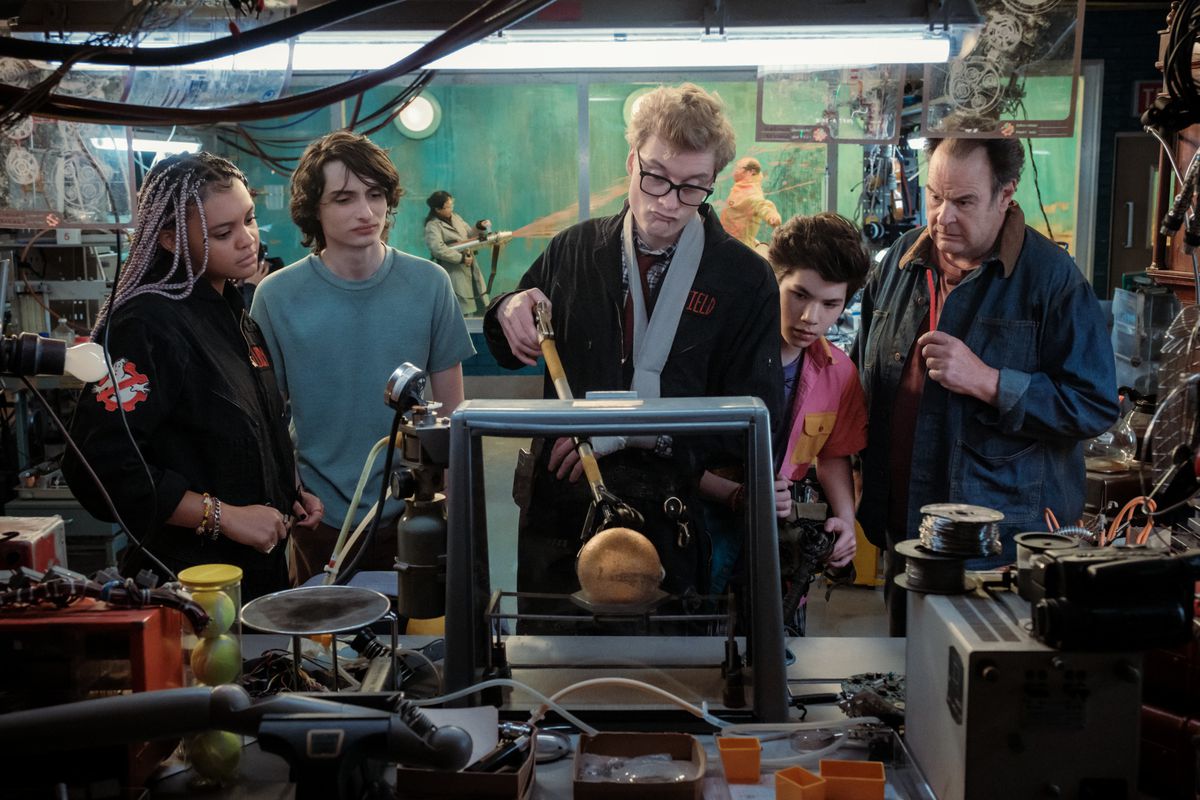 Un homme à lunettes montre aux chasseurs de fantômes un orbe doré tandis que quelqu'un tire du slime sur un scientifique en arrière-plan dans Ghostbusters : Frozen Empire.