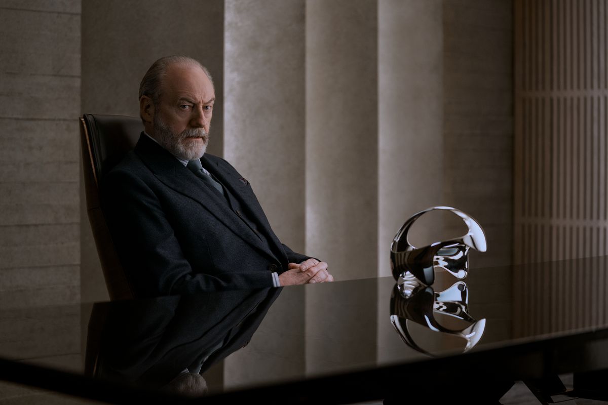 Thomas Wade (Liam Cunningham) assis à un bureau avec un casque futuriste sur une table.