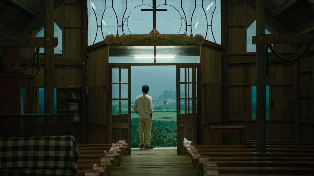 Un homme debout à l’entrée d’une église avec un vaste champ d’arbres et d’herbe au loin.