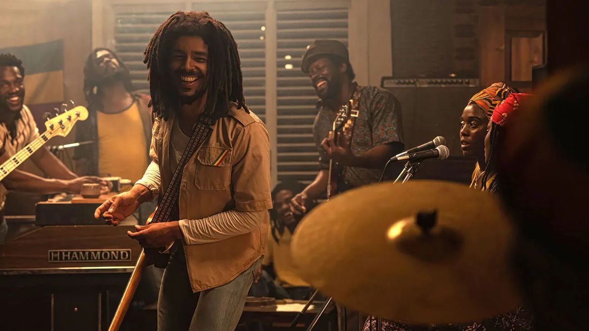 Un homme souriant avec des dreadlocks debout à côté d'un groupe de musiciens qui jouent.