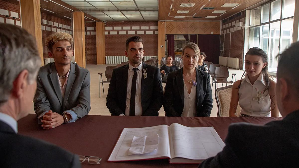 Deux hommes en costume et deux femmes en tenue formelle assis en face de deux messieurs plus âgés à une table avec un plus grand livre entre les deux.