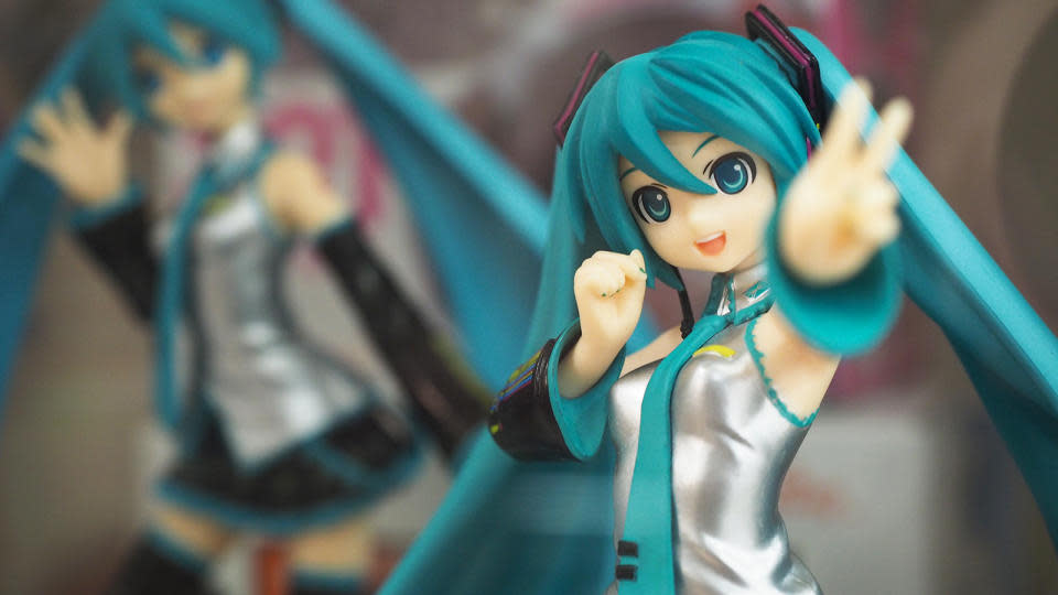 Gros plan d'une figurine Hatsune Miku.  Elle agite un signe de paix devant la caméra tandis qu'un deuxième personnage dans une pose différente est assis (flou) derrière.