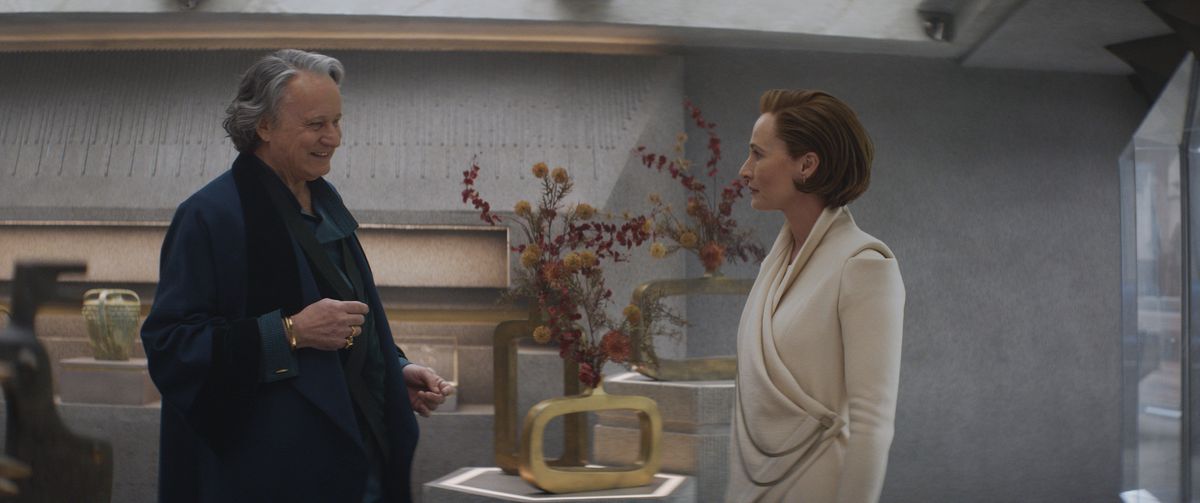 Luthen parle à Mon Mothma dans un magasin, avec lui debout devant elle.  Les fleurs sont dans des vases sur la table encadrés entre elles en arrière-plan