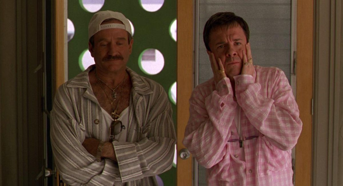 Robin Williams regarde vers le bas, les bras croisés.  Il porte une casquette de baseball blanche à l'envers, une chemise à col déboutonné et un maillot de corps blanc sur lequel pendent des lunettes de soleil.  Nathan Lane se tient à côté de lui, vêtu d'un haut rose, les mains couvrant son visage sous le choc.