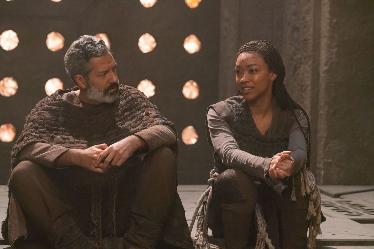 LR Alfredo Narciso dans le rôle d'Ohvahz et Sonequa Martin-Green dans le rôle de Michael Burnham dans Star Trek : Discovery.  Ils portent des vêtements extraterrestres fabriqués à la main et conversent calmement assis par terre dans une pièce en pierre. 