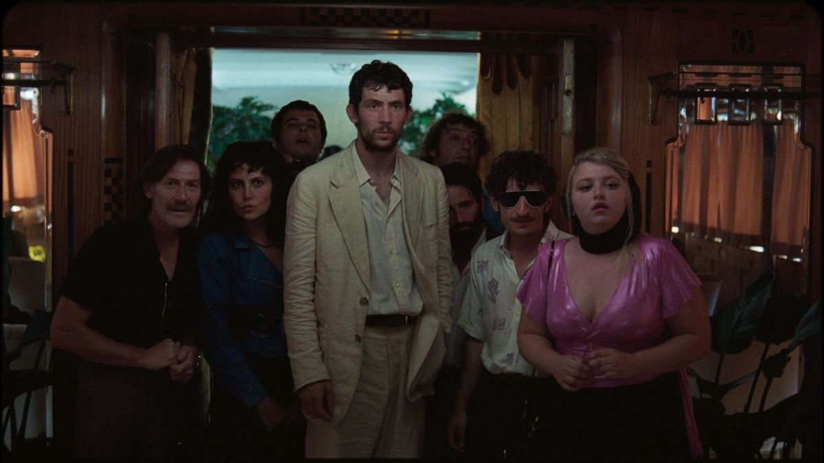 Un homme vêtu d'un costume blanc froissé avec une chemise boutonnée à col ouvert, entouré d'un groupe de personnes regardant quelque chose hors écran avec fascination.