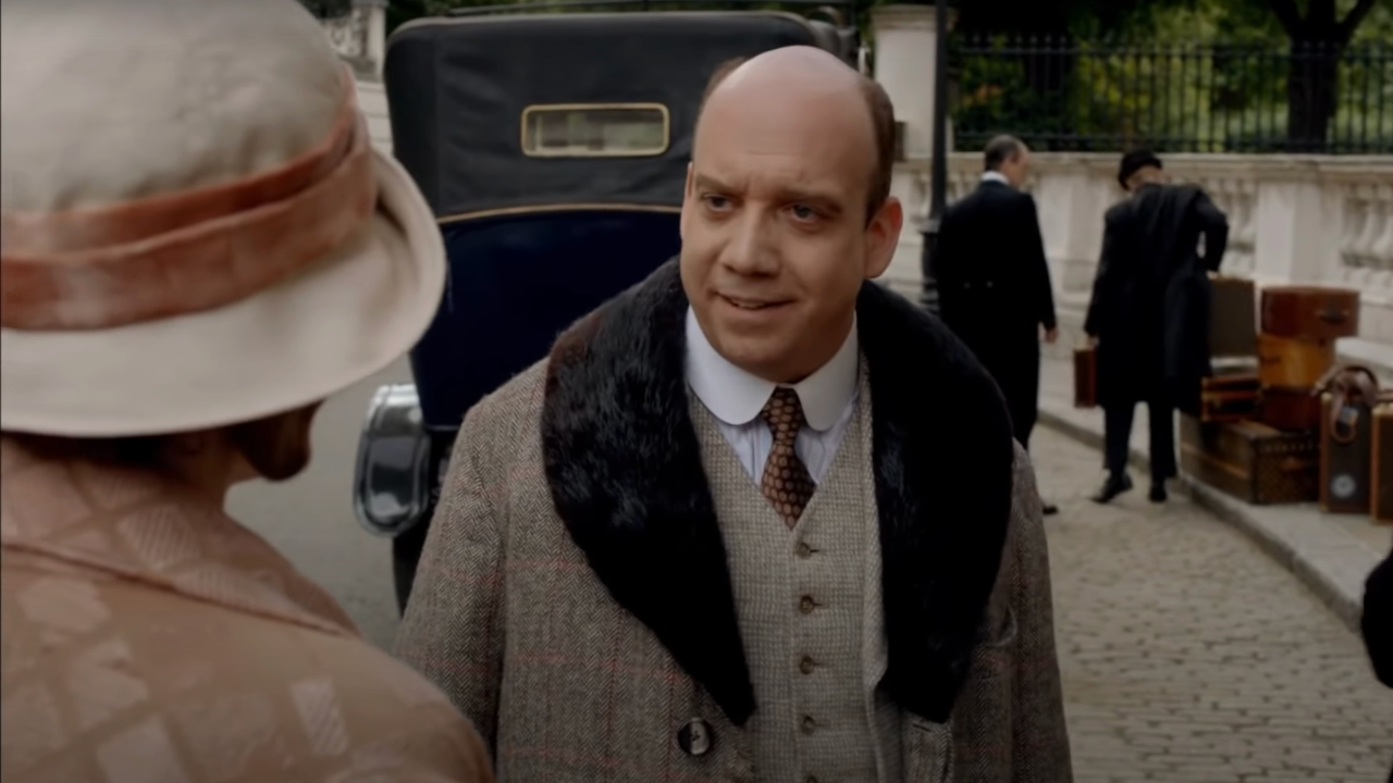 Paul Giamatti sourit à moitié alors qu'il se tient en costume d'époque devant Laura Carmichael à Downton Abbey.