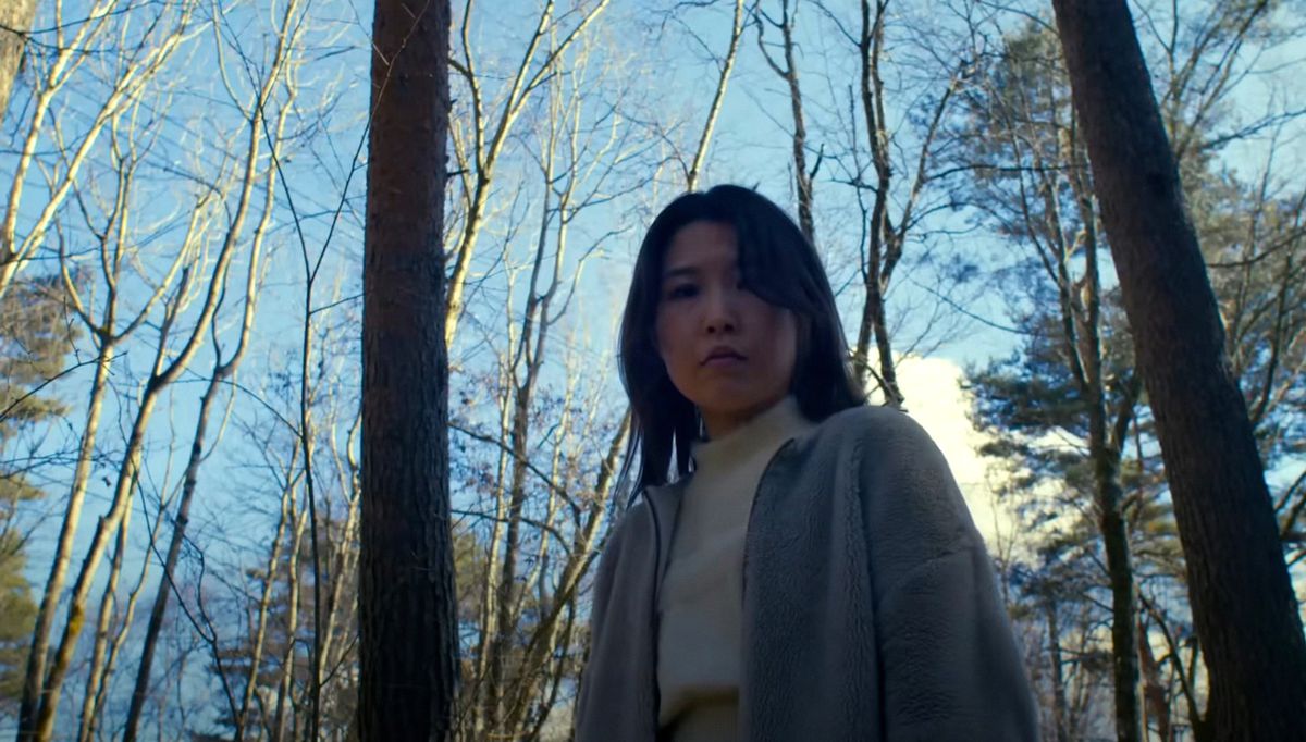 Mayuzumi (Ayaka Shibutani), une Japonaise vêtue d'une chemise blanche et d'un cardigan gris, se tient dans les bois, regardant la caméra vers le bas, dans Evil Does Not Exist de Ryûsuke Hamaguchi.