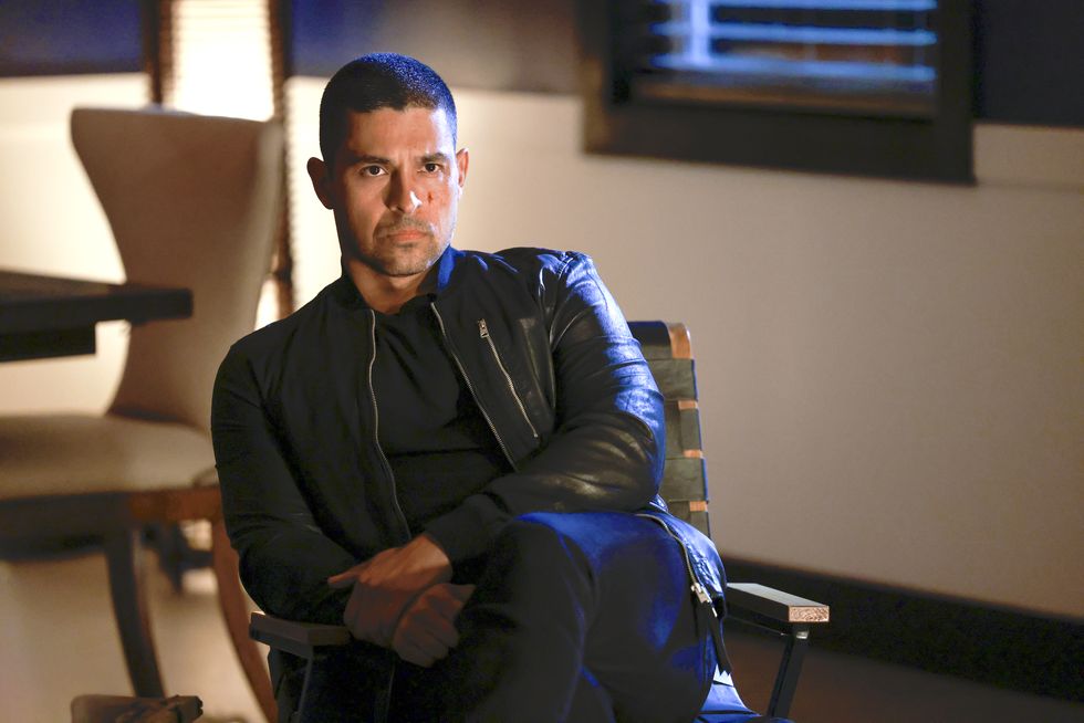 Wilmer Valderrama dans le rôle de Nick Torres dans la saison 20 de NCIS, un homme est assis sur une chaise et a l'air sévère, il porte un t-shirt noir, un jean et une veste en cuir.