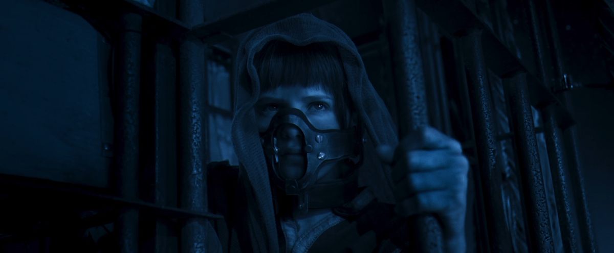 Furiosa enfant (Alyla Browne) est assise dans une cage la nuit, se tenant aux barreaux, portant une muselière et un drap sur la tête dans Furiosa de George Miller