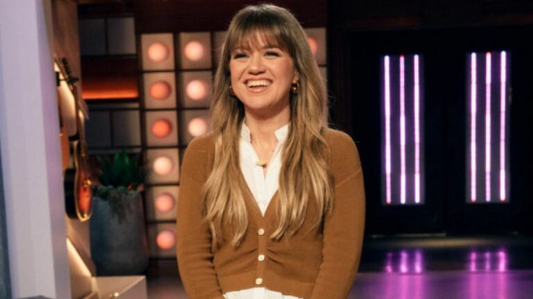 Kelly Clarkson, wearing a white shirt and brown cardigan, smiles at the crowd from the stage of The Kelly Clarkson Show.