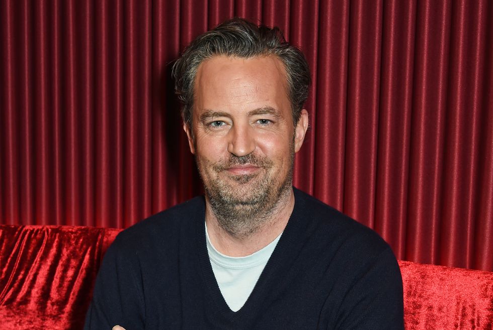 Matthew Perry est assis, les bras croisés, contre un canapé rouge avec un rideau rouge assorti derrière lui