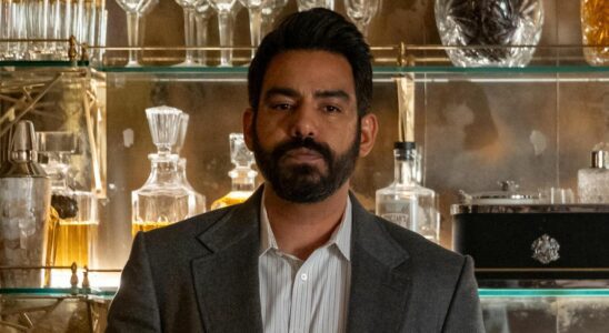 Sunil wearing a blazer and holding a glass of liquor while in front of a large bar full of alcohol bottles in Death and Other Details