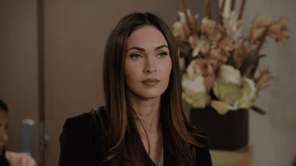 Megan Fox sits in a black blouse in front of a vase of flowers in a scene from New Girl