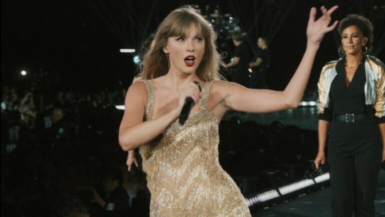 Taylor Swift holding her mic up and her free hand up while singing in the Fearless dress on The Eras Tour.