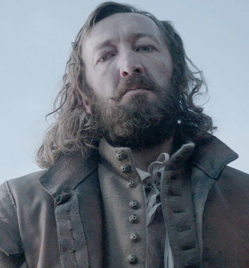 Ralph Ineson dans The Witch debout dans un grand manteau avec sa grande barbe au-dessus de la caméra