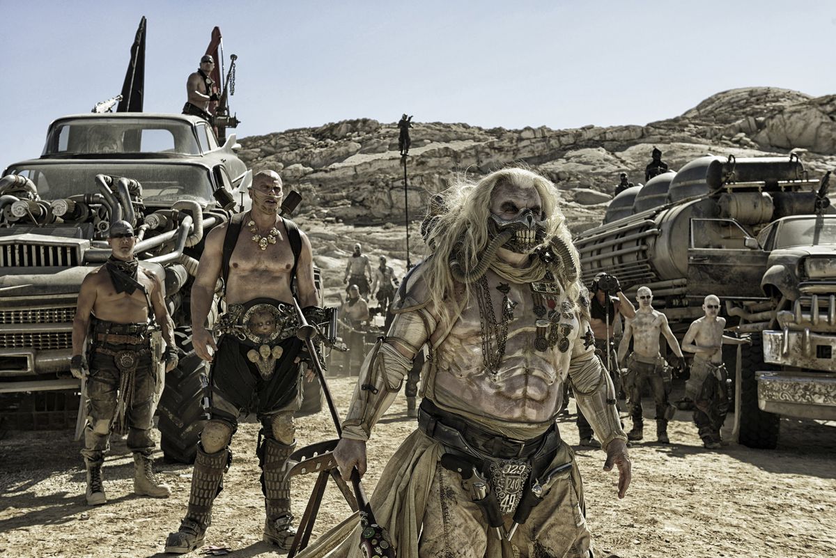 de gauche à droite : Nathan Jones dans le rôle de Rictus Erectus et Hugh Keays-Byrne dans le rôle d'Immortan Joe se tiennent entourés de War Boys et d'énormes véhicules dans Mad Max : Fury Road.