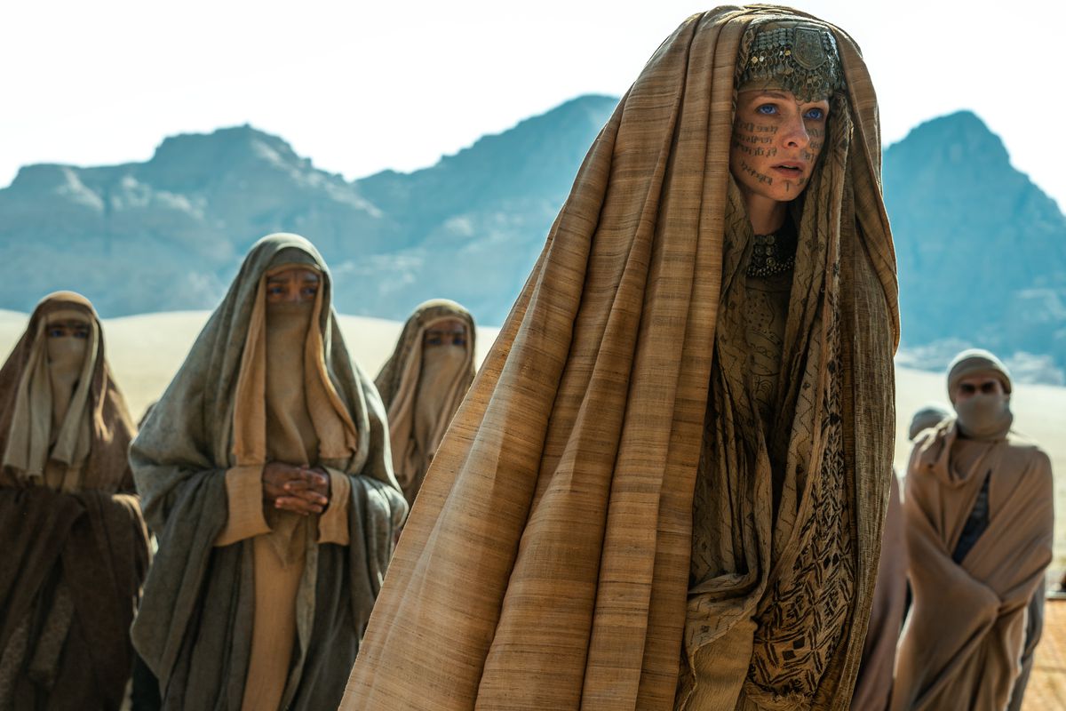 Lady Jessica (Rebecca Ferguson), encapuchonnée et avec des symboles écrits à l'encre sur son visage, se tient dans le désert, entourée de personnages vêtus de la même manière dans Dune de Denis Villeneuve : Deuxième partie