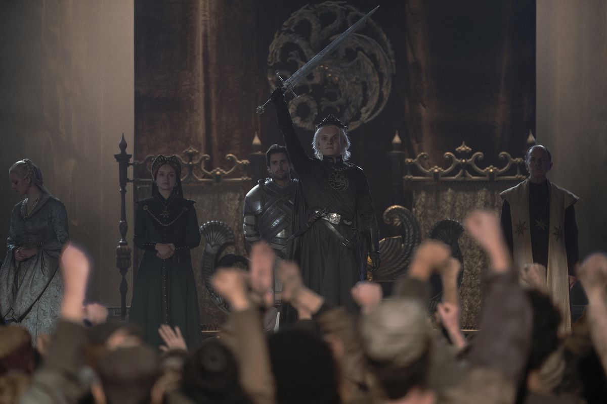 Le roi Aegon II (Tom Glynn-Carney) debout devant une foule de membres du public de Westeros en liesse, tenant triomphalement son épée en l'air