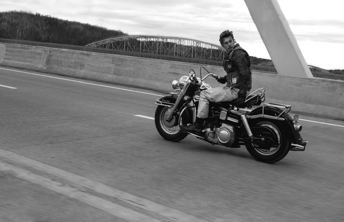 Austin Butler, en noir et blanc, conduit sa Harley-Davidson sur un pont d'une seule main, regardant par-dessus son épaule d'une manière très cool dans The Bikeriders