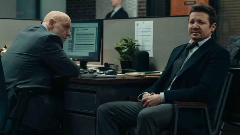 Hugh Dillon sitting in front of a computer looking at Jeremy Renner who is sitting by his desk facing forward.