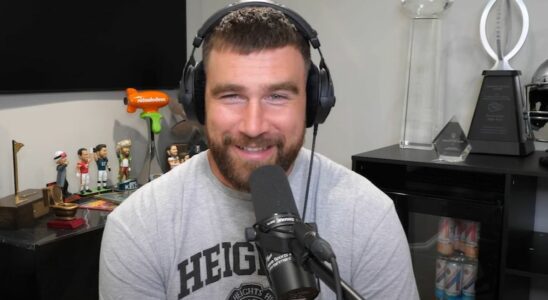 Travis Kelce smiling while wearing headphones and sitting behind a microphone during an episode of New Heights.