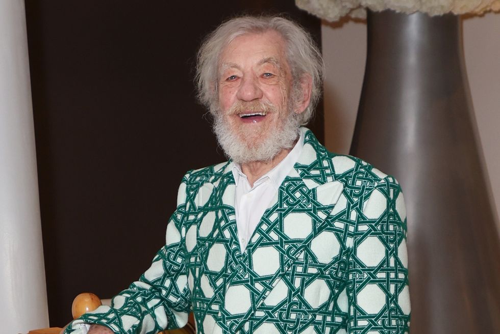 ian mckellen sourit alors qu'il se tient dans le hall d'un hôtel, le bras appuyé sur une pièce d'échecs géante