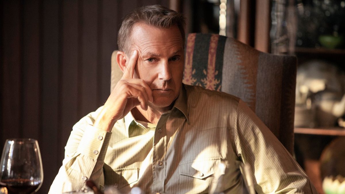 Kevin Costner leaning against his hand while sitting at the dinner table in Yellowstone.