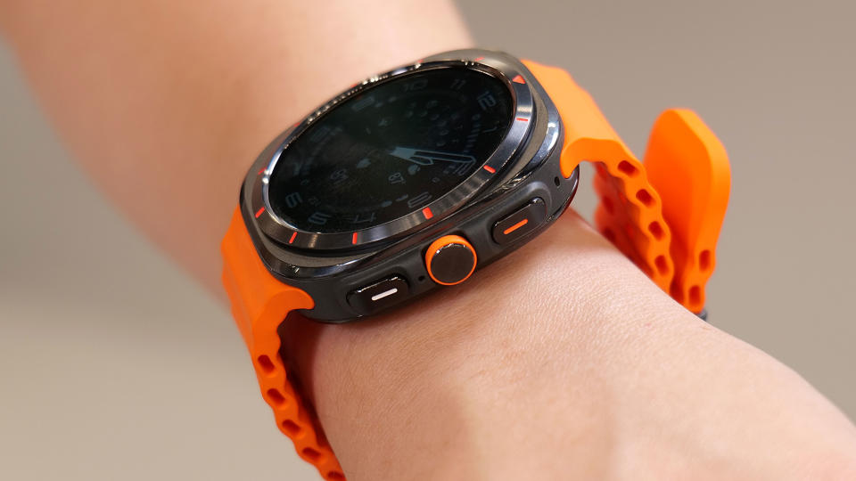 Vue latérale de la Samsung Galaxy Watch Ultra sur un poignet en l'air, montrant les trois boutons sur son bord droit. Le haut est un bouton rectangulaire noir avec une ligne orange à l'intérieur, tandis que le milieu est un cadran rond avec un contour orange. Le dernier est un rectangle noir avec une ligne blanche à l'intérieur.