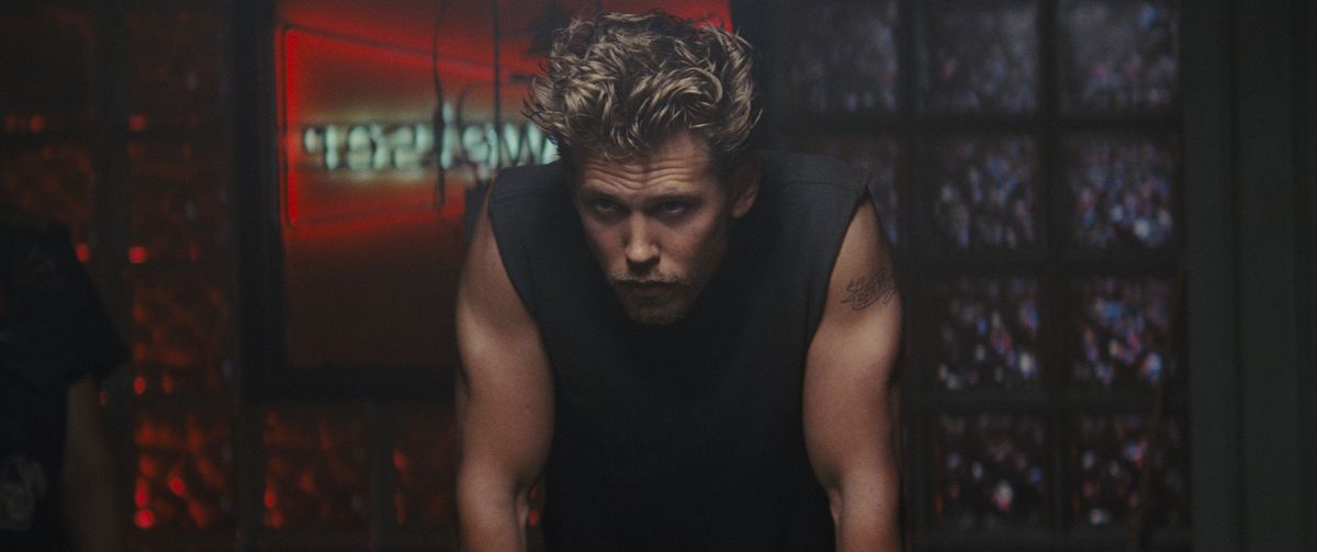 Austin Butler, les cheveux ébouriffés, portant un haut noir sans manches, se penche en avant d'une manière maussade dans The Bikeriders