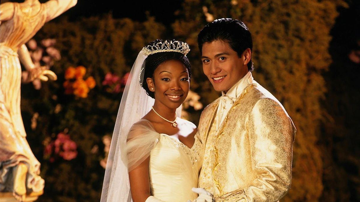 Une femme en robe blanche et diadème (Brandy Norwood) et un homme en costume blanc (Paolo Montalban) sourient devant un fond de fleurs et de statues.