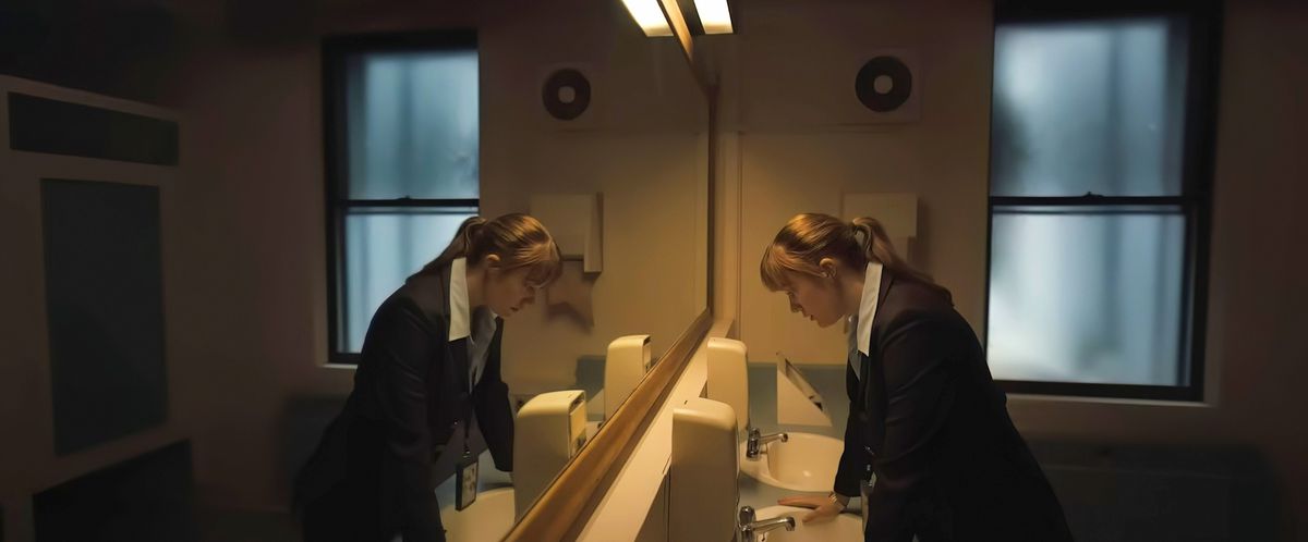 L'agent du FBI Lee Harker (Maika Monroe) se tient dans une grande salle de bain devant une rangée de lavabos et regarde vers le bas, reflété dans un miroir sur toute la longueur du mur, dans Longlegs d'Oz Perkins