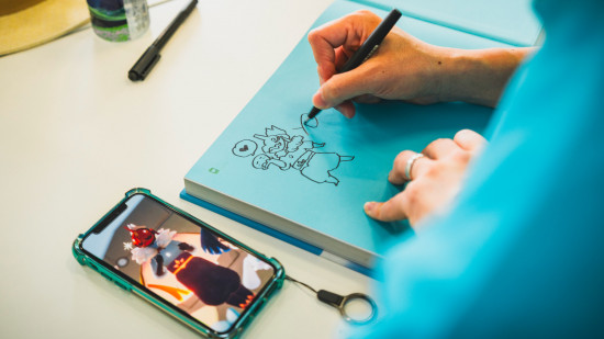 Ciel : communauté Enfants de la Lumière : une photographie par-dessus l'épaule d'un membre de l'équipe artistique signant un livre d'art, utilisant une photo du personnage d'un fan comme référence pour un gribouillage sur la couverture