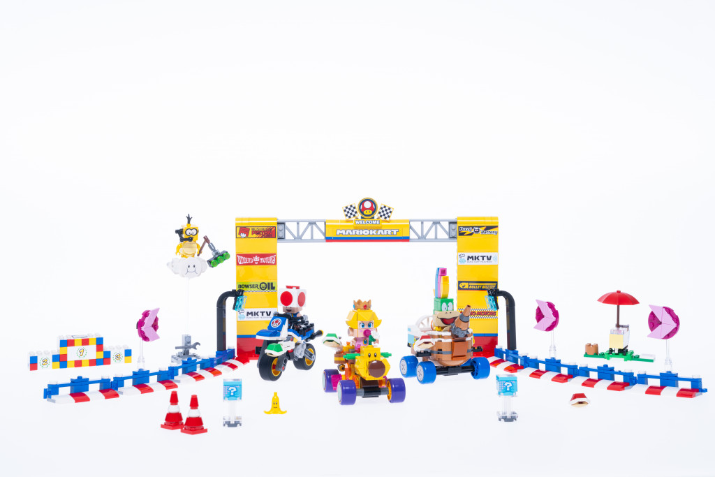 Images promotionnelles officielles de l'ensemble LEGO Mario Kart Baby Princess Peach et Grand Prix. On y voit une série de karts et de personnages construits en briques LEGO, dont Lemmy, un crapaud, Baby Peach et Lakitu. Les chariots et les personnages posent près de la ligne de départ d'un circuit de course.