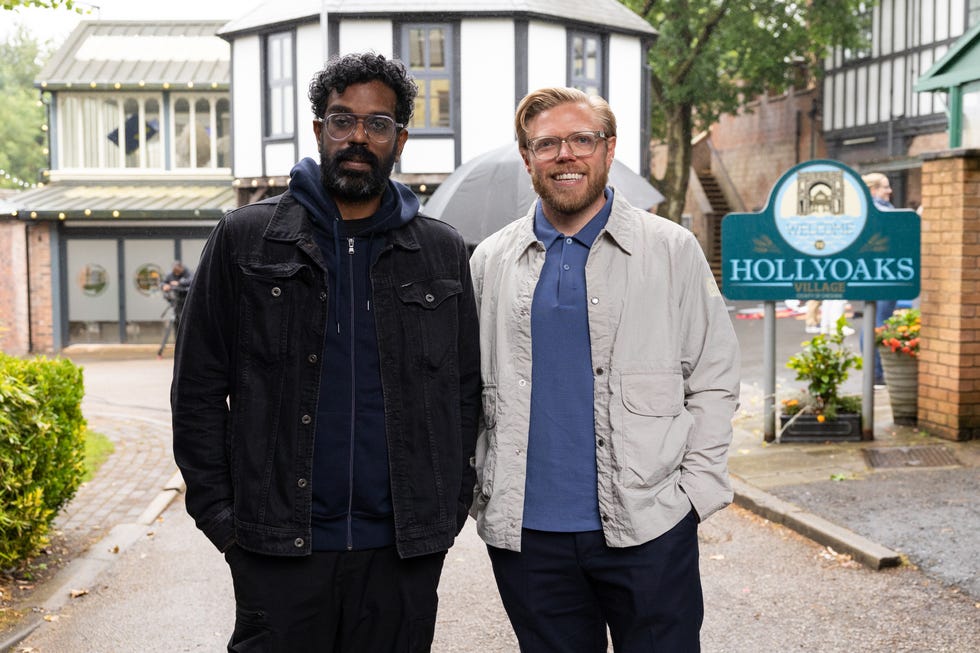 Romesh Ranganathan et Rob Beckett sourient devant le panneau du village de Hollyoaks