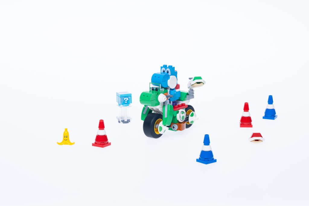 Images promotionnelles officielles de l'ensemble LEGO Mario Kart Yoshi Bike. On y voit un Yoshi bleu construit en briques LEGO sur l'emblématique moto Yoshi verte. Autour d'eux se trouvent des cônes et une peau de banane.