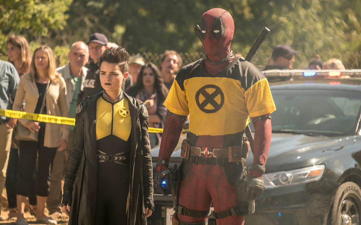 Deadpool (Ryan Reynolds, en costume, avec une version coupée d'un maillot de football jaune portant le symbole X-Man porté sur le dessus) se tient à côté de Negasonic Teenage Warhead (Brianna Hildebrand) devant un cordon de police et une foule de badauds dans Deadpool 2