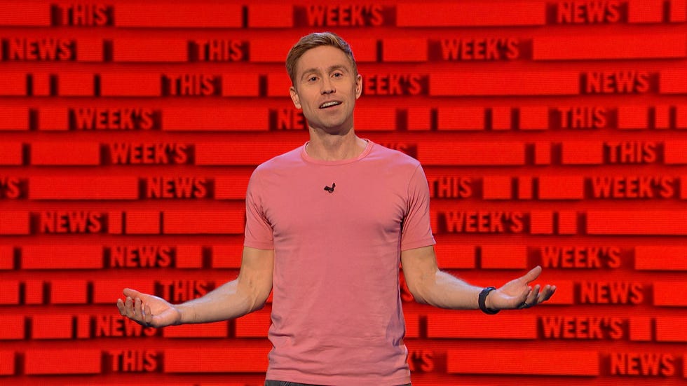 Russell Howard se tient devant l'écran avec les mots « les nouvelles de la semaine » répétés dessus alors qu'il présente l'heure de Russell Howard