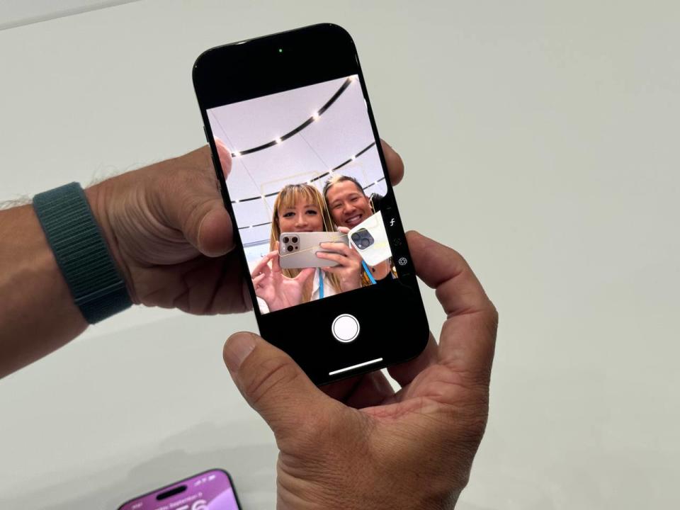 L'iPhone 16 Pro avec la caméra selfie allumée et deux personnes faisant face au téléphone. Le doigt d'une personne est posé sur le nouveau bouton de commande de l'appareil photo en bas à droite.