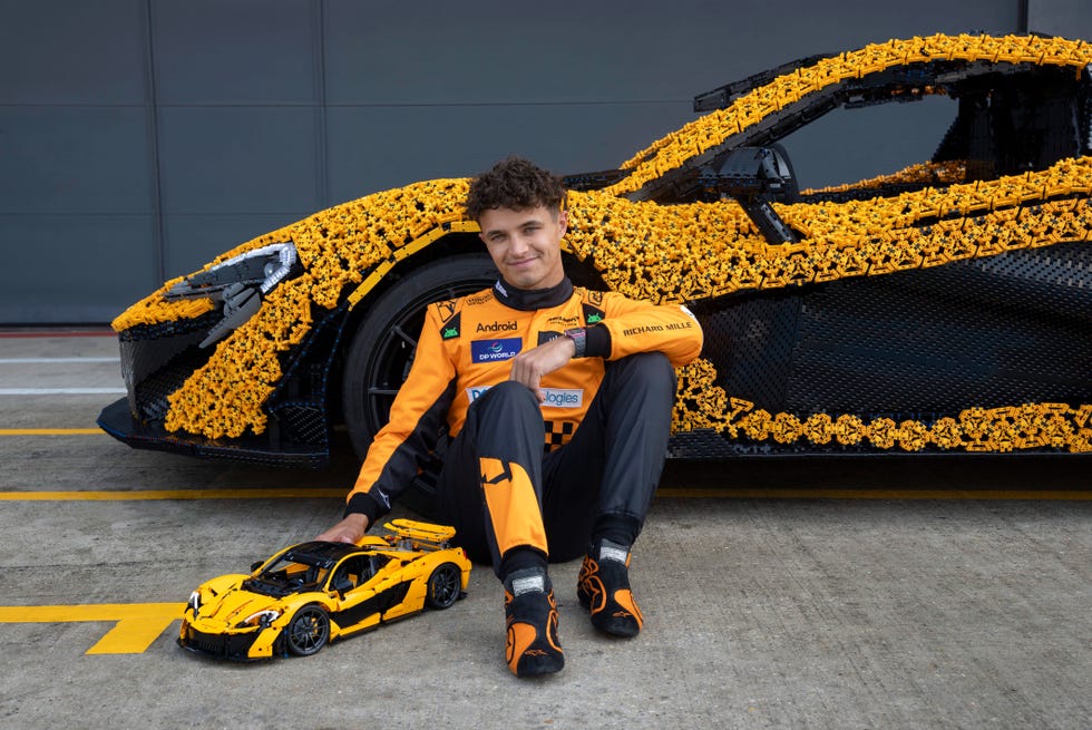 Lando Norris place sa main droite sur le modèle Lego Technic McLaren P1 alors qu'il est assis sur le sol devant la version grandeur nature