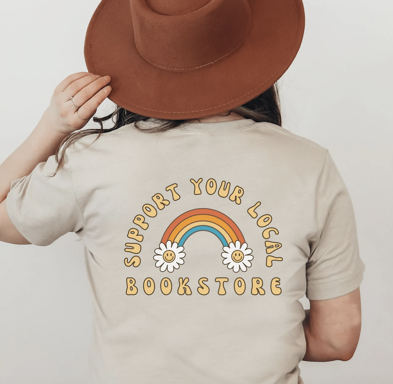 Image de dos de quelqu'un portant un t-shirt taupe avec un arc-en-ciel et la phrase "soutenez votre librairie locale" au dos de la chemise