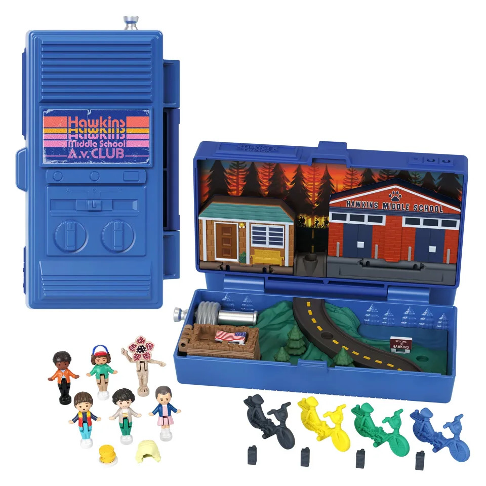 Le coffret Polly Pocket de Stranger Things et ses figurines sont présentés sur un fond blanc. Il comprend un compact bleu en forme de talkie-walkie, présenté fermé à gauche et ouvert à droite. Le côté ouvert contient une représentation de Hawkins, y compris le collège. Il y a également des figurines pour Eleven, Mike, Will, Lucas, Dustin et un Demogorgon, ainsi que des mini talkies-walkies et des vélos