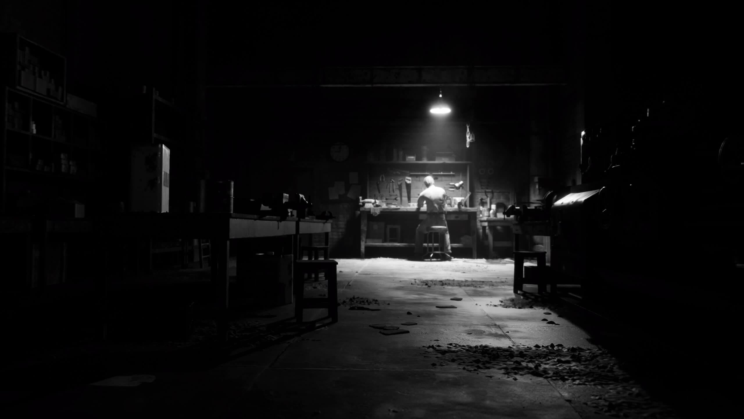 Un homme seul est assis dans son atelier. Un faisceau lumineux est dirigé vers lui pour ajouter une touche visuelle plus menaçante. L'ensemble de la prise de vue est recouvert d'un filtre noir et blanc.